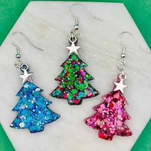 Puede incluir: Tres pendientes colgantes con forma de árbol de Navidad. Cada pendiente presenta un árbol lleno de purpurina en azul, verde y rosa, coronado por una estrella plateada. Los pendientes cuelgan de ganchos plateados.