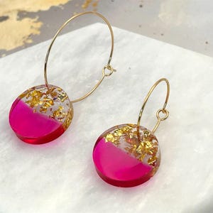 May include: A pair of hoop earrings featuring circular resin charms. Each charm is divided into two sections: one half is a vibrant hot pink, while the other half is clear and filled with metallic gold leaf flakes. The charms hang from thin gold-tone metal hoops.