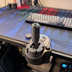 May include: A black and silver Logitech gaming shifter is mounted on a desk. The shifter has a textured handle and a base with the Logitech logo. A keyboard and desk mat are in the background.