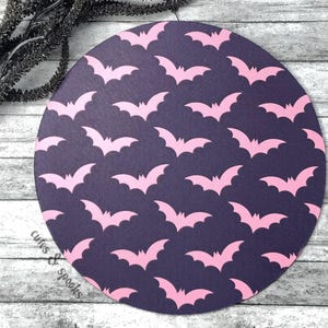 May include: A round, dark purple coaster with a repeating pattern of pink bats. The coaster is perfect for adding a touch of spooky fun to your home decor.