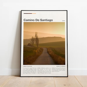 May include: A framed print depicting the Camino de Santiago in Spain. The image shows a path winding through fields, with text that reads "Camino De Santiago" and "Spain". The colour palette is warm, with tones of brown, orange, and green, evoking a sense of travel.