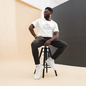 May include: White t-shirt with a black line drawing of a vehicle. The shirt is worn by a person wearing black jeans and white sneakers. The person is sitting on a black stool against a beige and black backdrop.