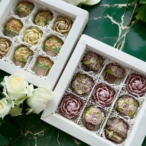 Könnte beinhalten: Zwei weiße Schachteln gefüllt mit dekorativen Pralinen in Form von Blumen. Die Pralinen sind in verschiedenen Farben, darunter Weiß, Gelb und Lila. Die Schachteln stehen auf einer grünen Marmoroberfläche, mit weißen Rosen.