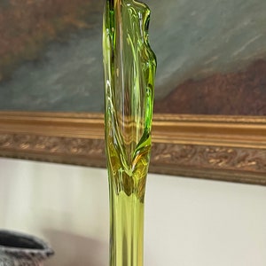 May include: A tall, slender, lime green glass vase with a unique, organic shape. The vase has a flared top and a smooth, reflective surface. The background features a painting with a gold frame.