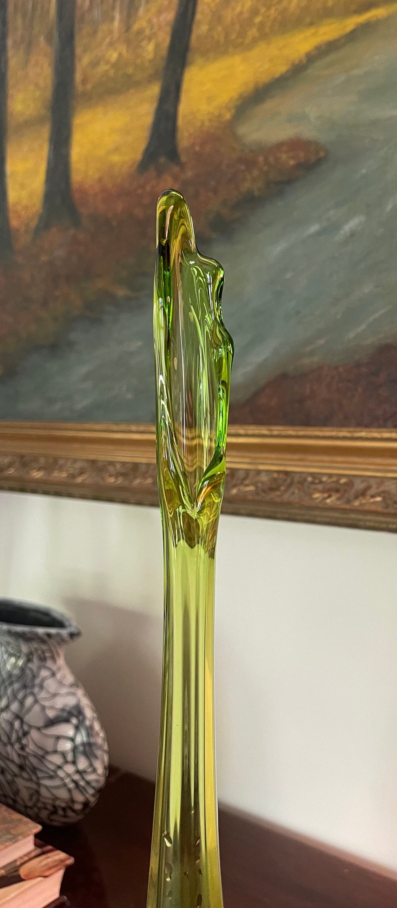 May include: A tall, slender, lime green glass vase with a unique, organic shape. The vase has a flared top and a smooth, reflective surface. The background features a painting with a gold frame.