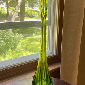 May include: A tall, slender, lime green glass vase with a flared base and a hexagonal foot. The vase has a long, elegant neck that tapers to a pointed top. The vase is displayed on a dark wooden table near a window.