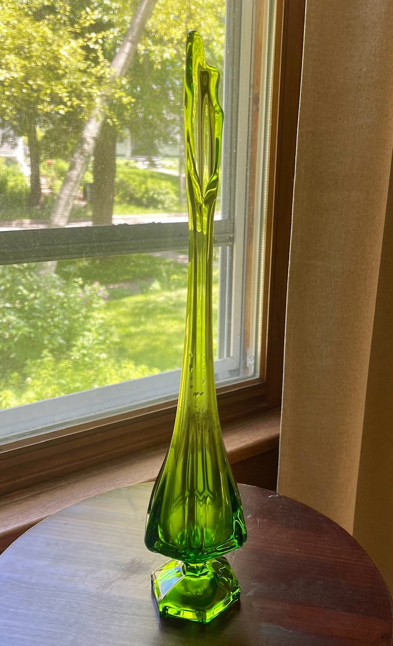 May include: A tall, slender, lime green glass vase with a flared base and a hexagonal foot. The vase has a long, elegant neck that tapers to a pointed top. The vase is displayed on a dark wooden table near a window.