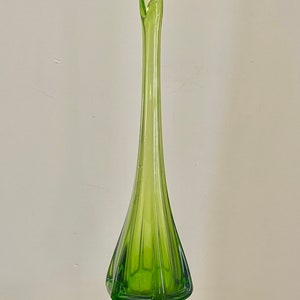 May include: Tall, green glass vase with a flared top and a geometric base. The vase has a slender, elongated body with vertical ridges, and it stands on a hexagonal base. The vase is a vibrant shade of green.