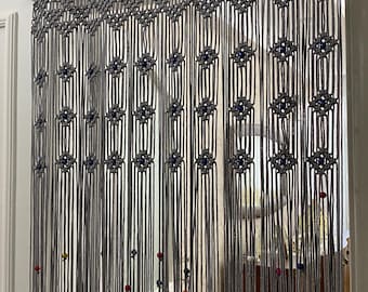 May include: Grey macrame curtain with colourful beads hanging from a wooden rod. The curtain is made of knotted cords and has a geometric pattern.