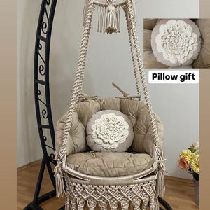 May include: A beige macrame hanging chair with a plush cushion and a matching pillow. The chair is suspended from a black metal stand. "Pillow gift" is written below the pillow.