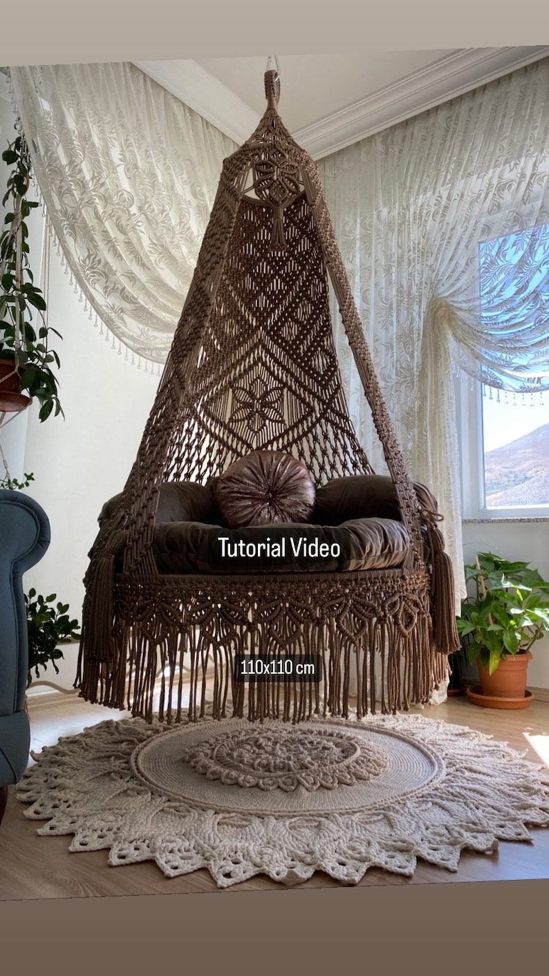 May include: A brown macrame hanging chair with a fringe border and a plush cushion. The chair is 110x110 cm and is hanging in a room with white curtains. The text "Tutorial Video" is visible on the chair.