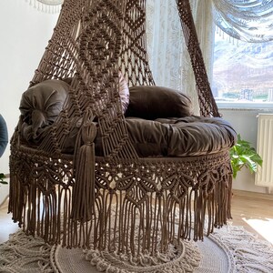 May include: A brown macrame hanging chair with a fringe border and a thick cushion. The chair is hanging from the ceiling and is in a room with white curtains and a rug.