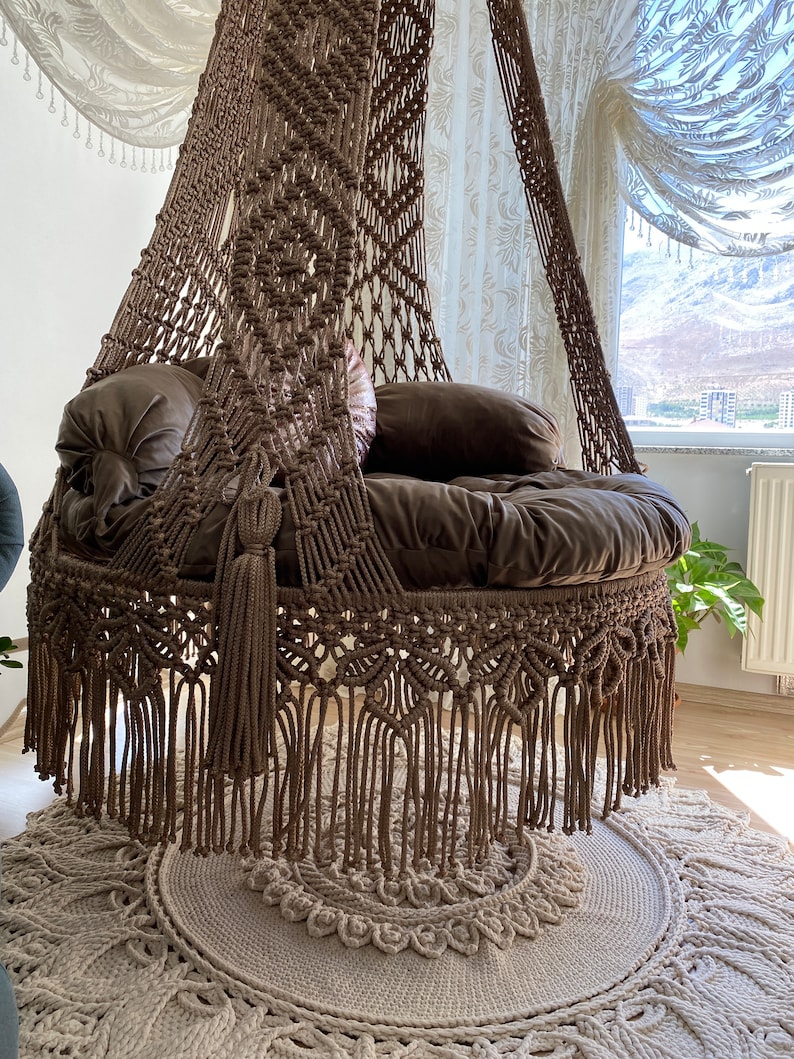 May include: A brown macrame hanging chair with a fringe border and a thick cushion. The chair is hanging from the ceiling and is in a room with white curtains and a rug.