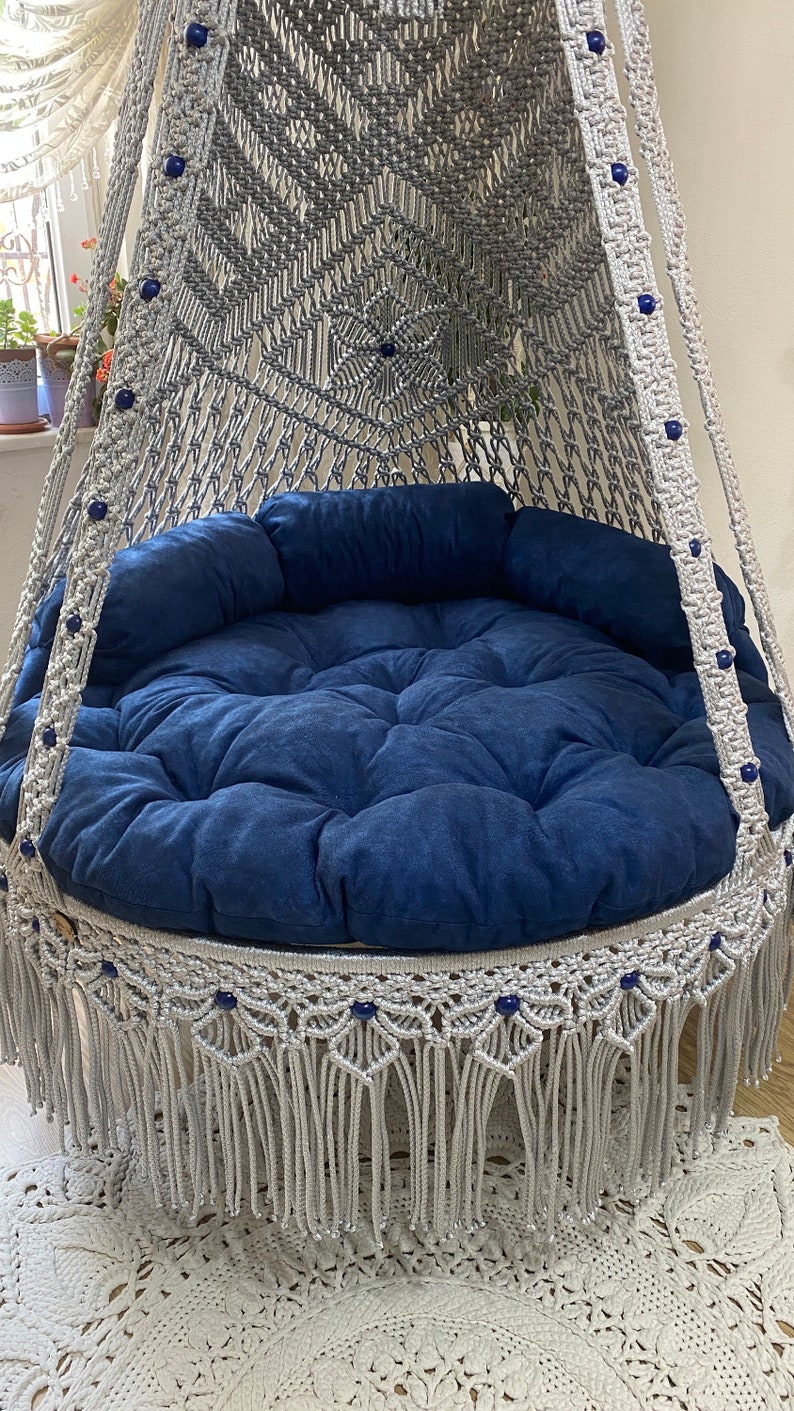 May include: A gray macrame hanging chair with a blue cushion and fringe trim. The chair is decorated with blue beads.