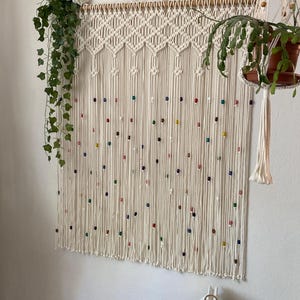 May include: A white macrame wall hanging with colorful beads. The hanging is made with a knotting technique and has a diamond pattern at the top.