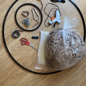 May include: A collection of craft supplies for a macrame project, including a black metal hoop, a roll of beige rope, a measuring tape, a glue gun, a pair of scissors, zip ties, and a carabiner.