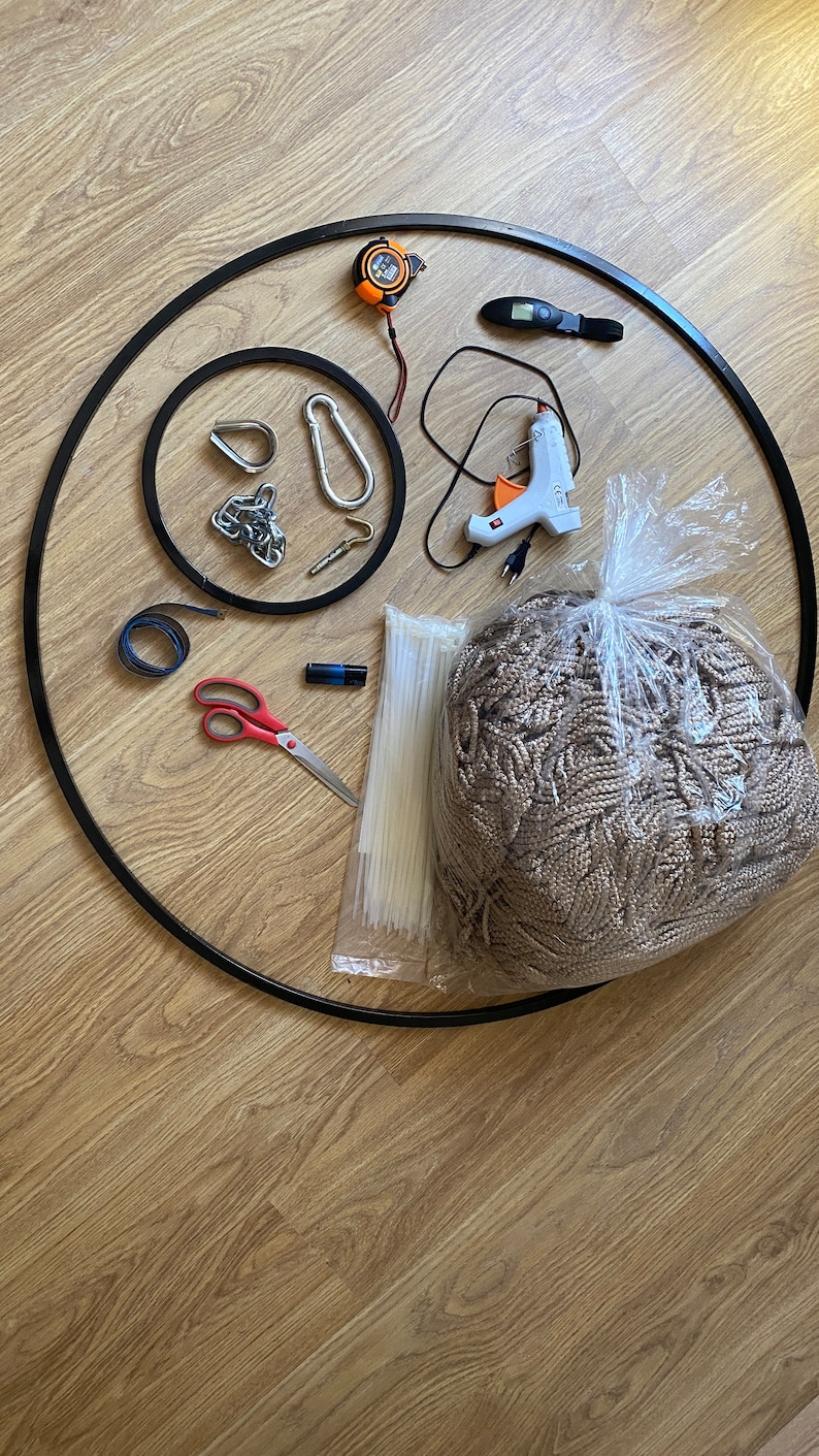 May include: A collection of craft supplies for a macrame project, including a black metal hoop, a roll of beige rope, a measuring tape, a glue gun, a pair of scissors, zip ties, and a carabiner.