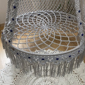 May include: A gray macrame hanging chair with blue beads and fringe. The chair is hanging over a white crocheted rug.