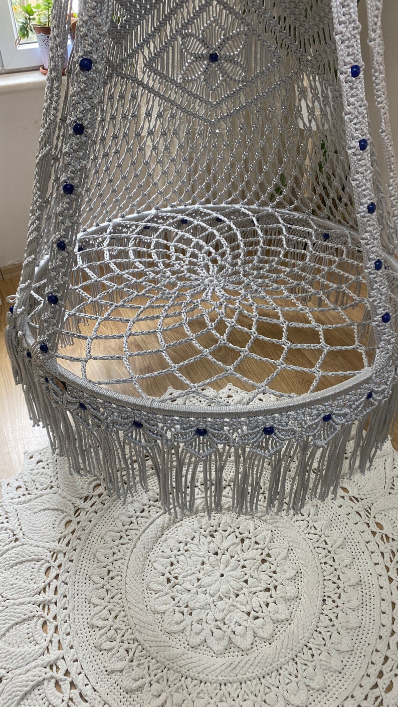May include: A gray macrame hanging chair with blue beads and fringe. The chair is hanging over a white crocheted rug.