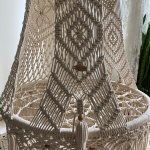 May include: A white macrame baby crib canopy with a circular base and wooden beads. The canopy is made with a diamond pattern and has a tassel hanging from the bottom.
