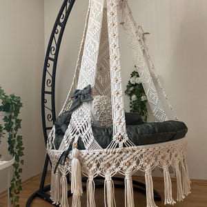 May include: A cream-colored macrame swing chair with a black metal frame. The chair features a woven design, wooden beads, and a fringed bottom. Dark gray cushions and pillows provide seating. A decorative, bohemian-style hanging chair.