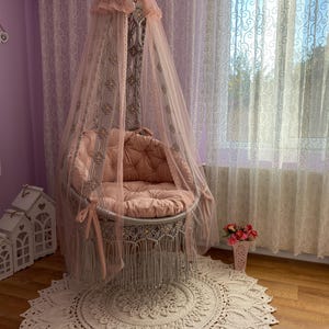 May include: A white macrame hanging chair with a pink canopy and a pink cushion. The chair is hanging in a room with white curtains and a white rug.