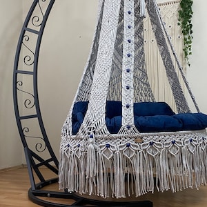 May include: A white macrame hanging chair with a blue cushion, suspended from a black metal stand. The chair has a fringe border and blue beads.