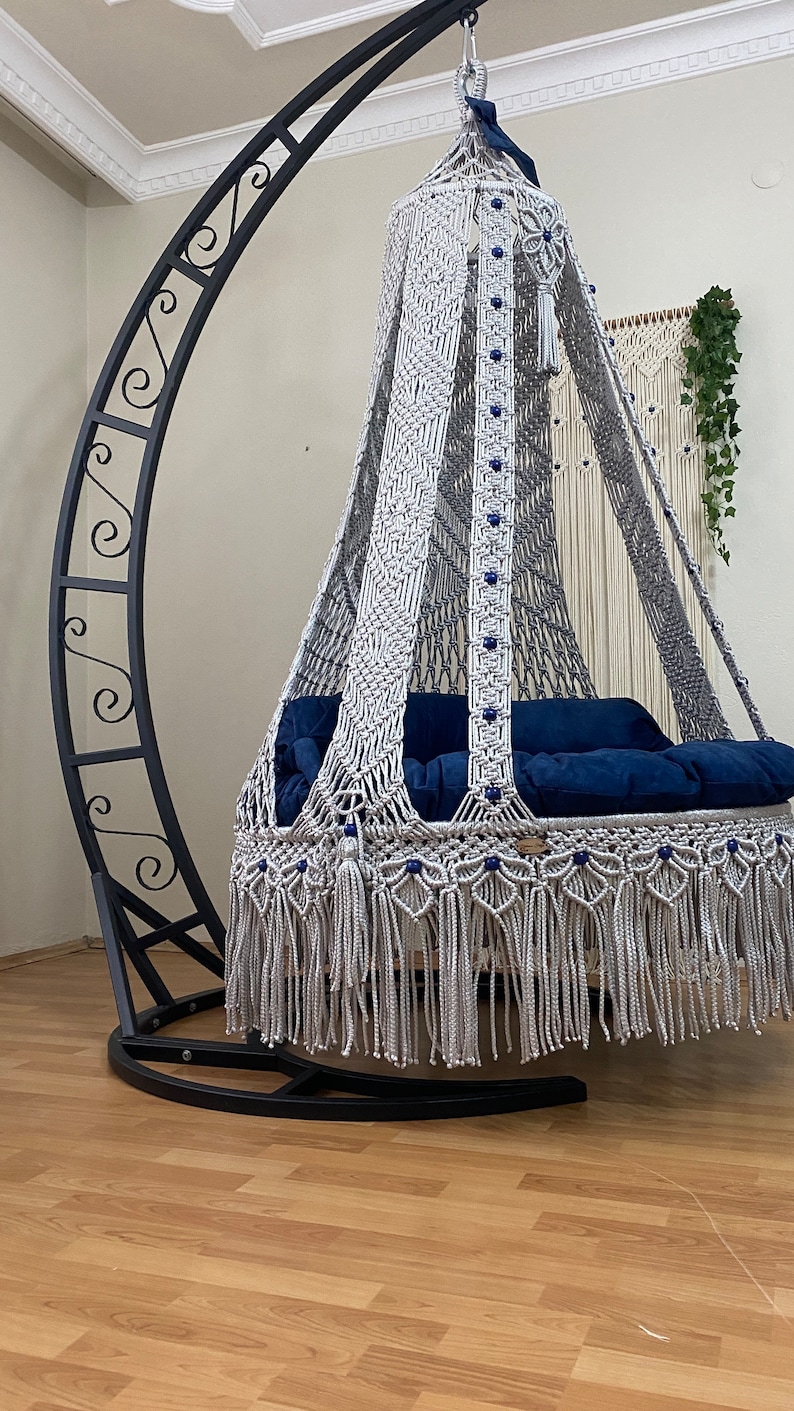 May include: A white macrame hanging chair with a blue cushion, suspended from a black metal stand. The chair has a fringe border and blue beads.