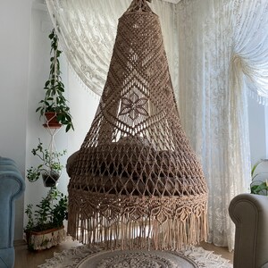 May include: A large, beige macrame hanging chair with a fringe border. The chair is suspended from the ceiling in a room with white walls and sheer curtains.