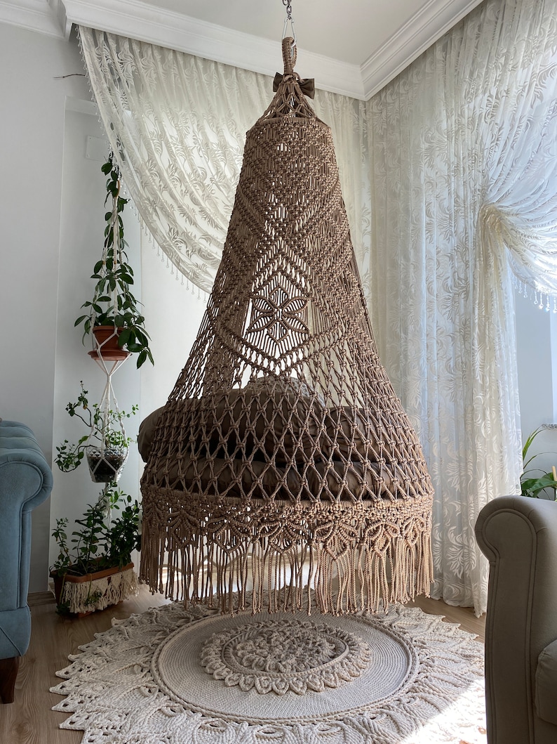 May include: A large, beige macrame hanging chair with a fringe border. The chair is suspended from the ceiling in a room with white walls and sheer curtains.