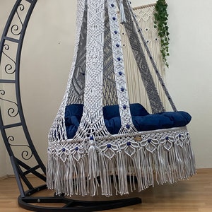 May include: A white macrame hanging chair with a blue cushion and a black metal stand. The chair has a fringe border and is decorated with blue beads.