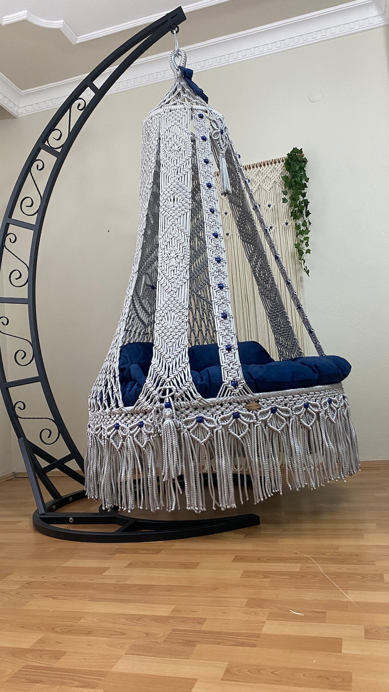May include: A white macrame hanging chair with a blue cushion and a black metal stand. The chair has a fringe border and is decorated with blue beads.