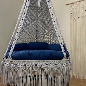 May include: A white macrame hanging chair with a blue cushion and blue beads. The chair is hanging from the ceiling and has a fringe around the bottom.