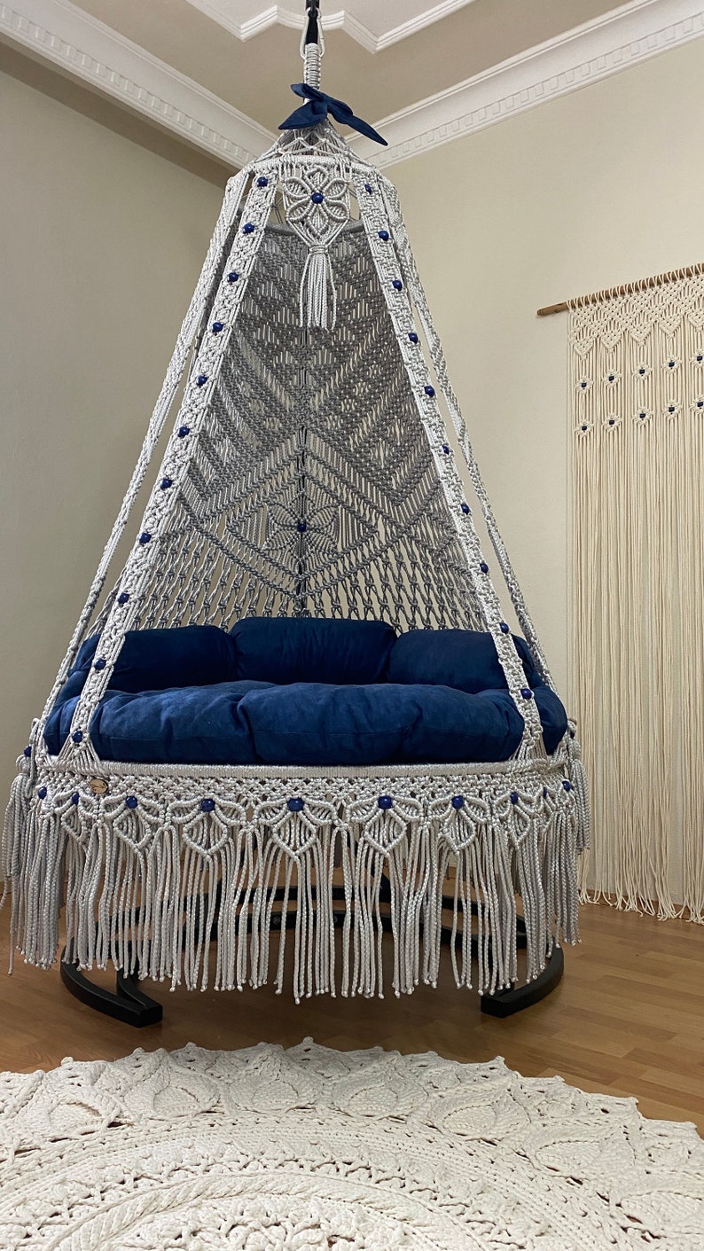May include: A white macrame hanging chair with a blue cushion and blue beads. The chair is hanging from the ceiling and has a fringe around the bottom.