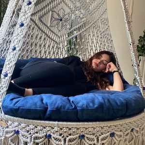 May include: A white macrame hammock chair with blue beads and a blue cushion. A person is lying in the hammock chair.