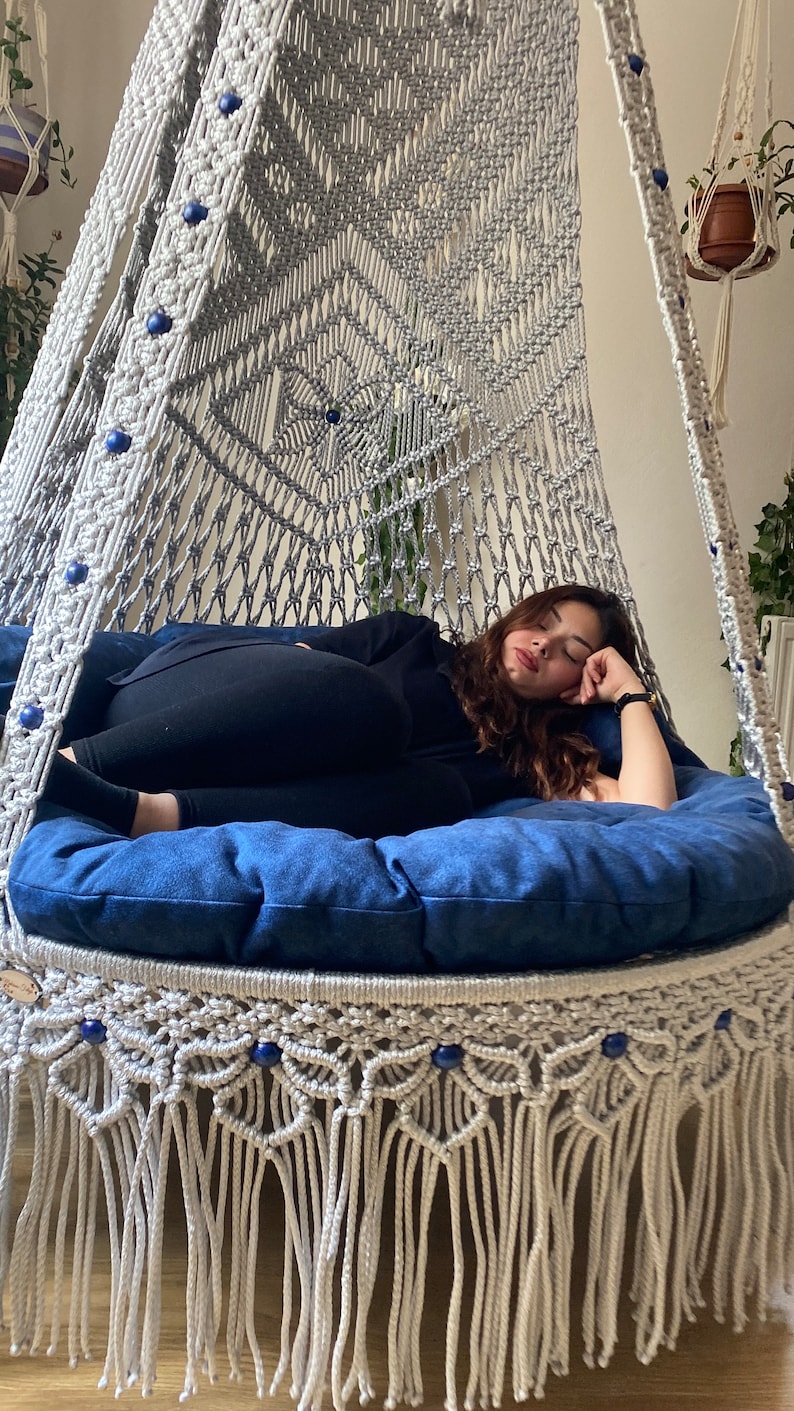 May include: A white macrame hammock chair with blue beads and a blue cushion. A person is lying in the hammock chair.