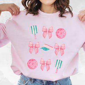 May include: Pink sweatshirt with a graphic design of pink bows, green and white paddle rackets, and pink pickleball balls.