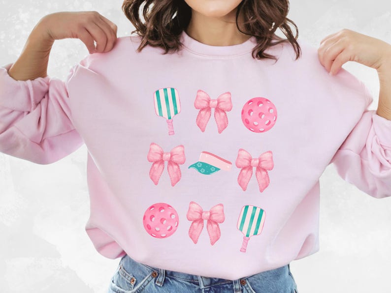 May include: Pink sweatshirt with a graphic design of pink bows, green and white paddle rackets, and pink pickleball balls.