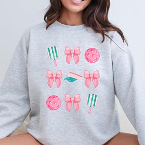 May include: A gray sweatshirt with a design of pink bows, green and white paddles, and pink and white pickleballs.