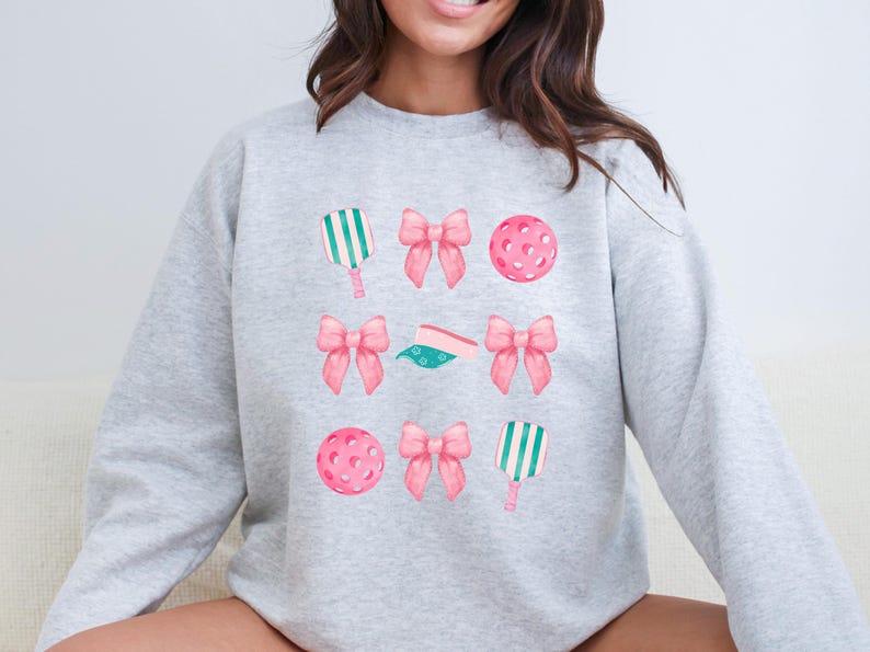 May include: A gray sweatshirt with a design of pink bows, green and white paddles, and pink and white pickleballs.