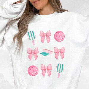 May include: White sweatshirt with a pink and green design featuring a pickleball paddle, pickleball, and bows.