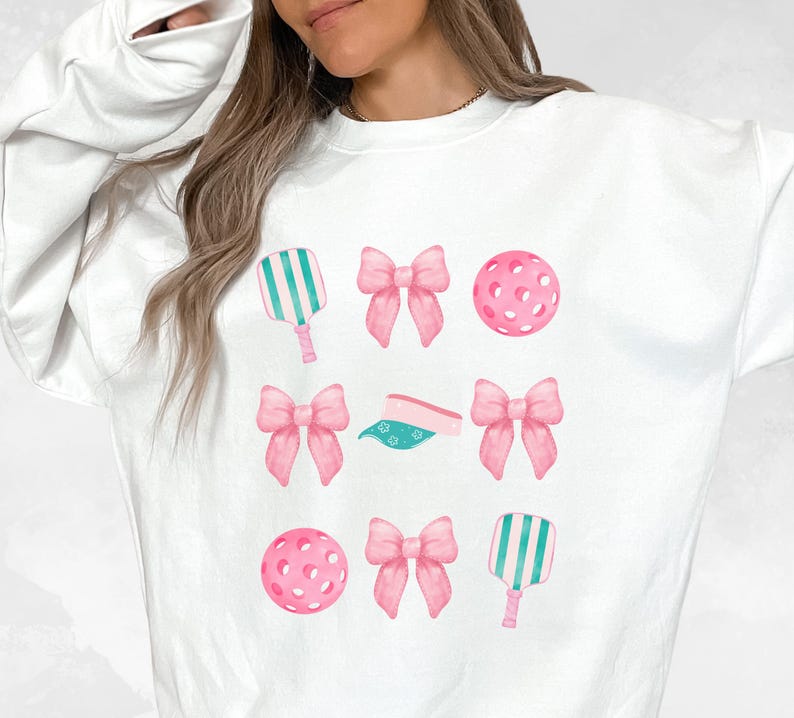 May include: White sweatshirt with a pink and green design featuring a pickleball paddle, pickleball, and bows.