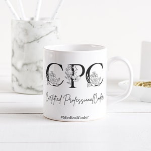 Op de afbeelding: Witte keramische mok met zwarte letters "CPC", versierd met bloemmotieven. De tekst "Certified Professional Coder" en de hashtag "#MedicalCoder" staan ook op de mok. Een marmeren pennenhouder en kantoorbenodigdheden zijn op de achtergrond.