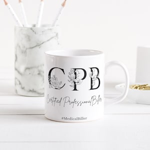 Op de afbeelding: Witte keramische mok met zwarte tekst die "CPB Certified Professional Biller #MedicalBiller" luidt. De letters zijn versierd met bloemmotieven. De mok staat op een wit houten oppervlak.