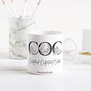 Op de afbeelding: Witte keramische mok met zwarte tekst die "COC Certified Outpatient Coder #MedicalCoder" luidt. De letters zijn versierd met bloemmotieven. De mok staat op een wit houten oppervlak.