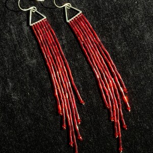 May include: A pair of red beaded earrings with silver-colored hooks. The earrings feature a triangular top with multiple strands of red beads cascading downwards. The beads have a slight sheen, and the earrings are set against a dark background.