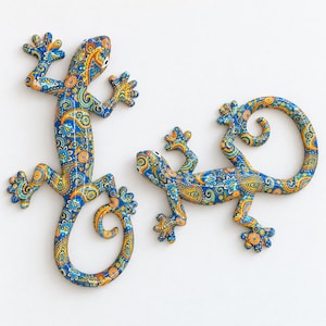 Könnte beinhalten: Zwei echsenförmige Wanddekorationen mit einem lebhaften Paisley-Muster in Blau, Gelb und Orange. Eine Echse ist vertikal mit eingerolltem Schwanz positioniert, während die andere horizontal ausgerichtet ist und einen kreisförmig geringelten Schwanz hat.