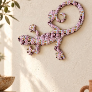 Könnte beinhalten: Eine Wandskulptur in Form eines Geckos mit einem detaillierten Mosaikmuster in Rosa-, Lila- und Orangetönen. Die Echsenfigur hat einen geschwungenen Schwanz und eine strukturierte Oberfläche mit einem sich wiederholenden kreisförmigen Muster.