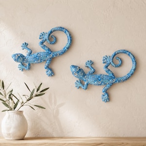 May include: Two wall-mounted lizard sculptures featuring a vibrant blue and white paisley pattern. Each lizard is depicted in a curved, climbing position with detailed toes and a curled tail, creating a decorative display on a neutral wall surface.
