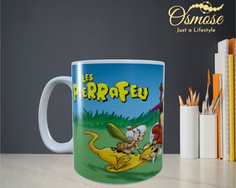 Puede incluir: Taza de cerámica blanca con una imagen de dibujos animados de un personaje montando un animal amarillo. El texto "LES PIERRAFIEU" está impreso en amarillo sobre un fondo azul.